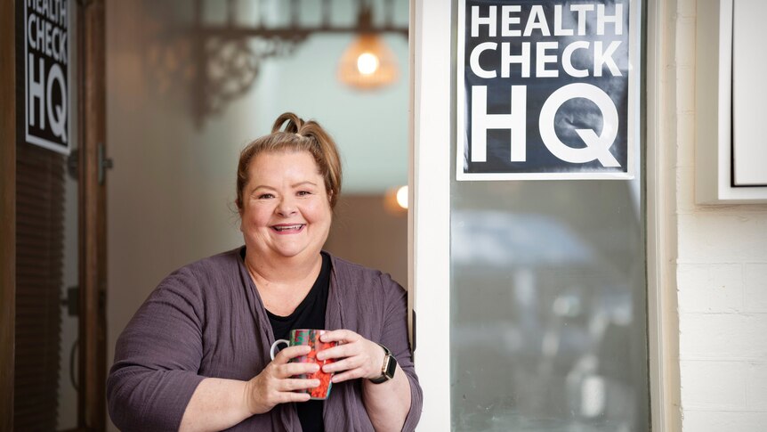 A smiling woman holds a coffee mug with both hands and leans against the jamb of a door, beside a sign saying Health Check HQ.