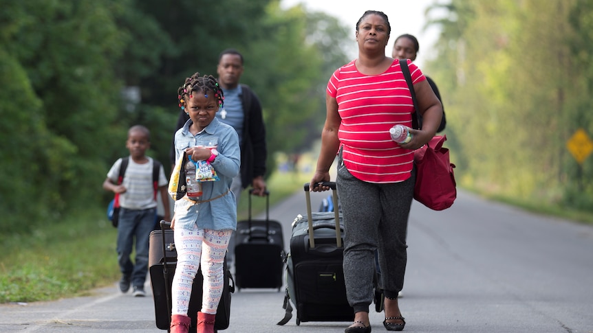 A family walk down a road with suitcases.