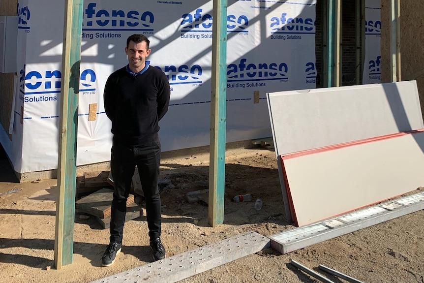 Man wearing dark jumper, black jeans and sneakers standing outside building site