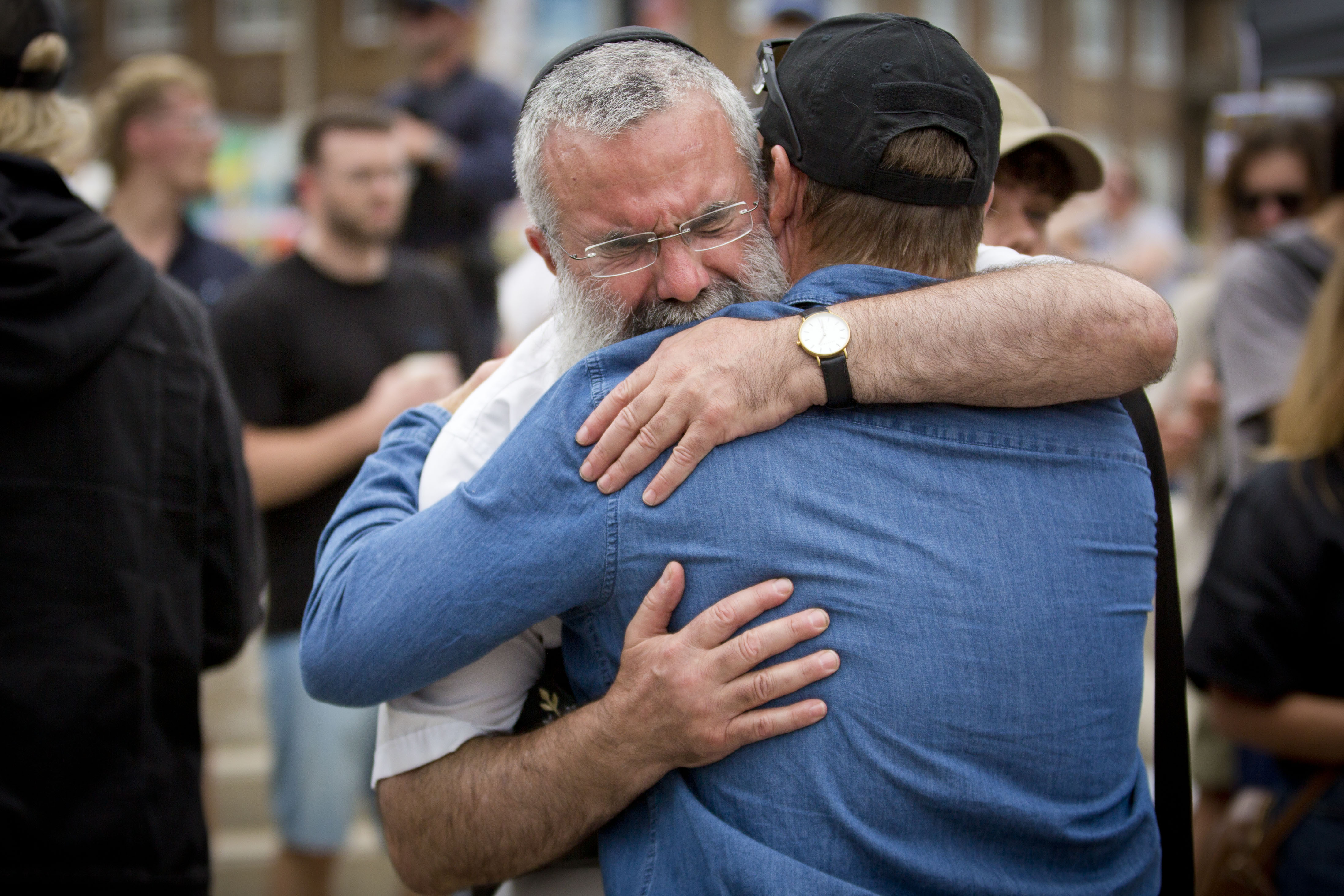 Dois homens, um deles chorando, se abraçam com força em um memorial ao ar livre