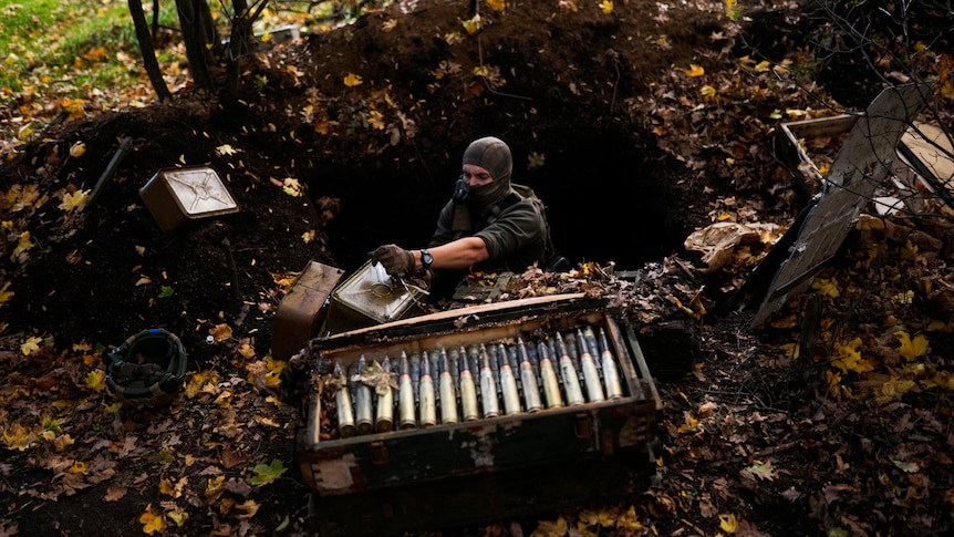 A man in camoflauge sits in a hole as he works to de-mine with various ammunition sits around him.