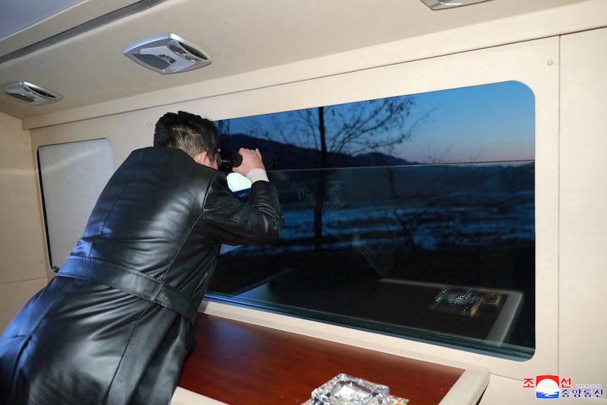 Man in leather coat using binoculars looks out a window.