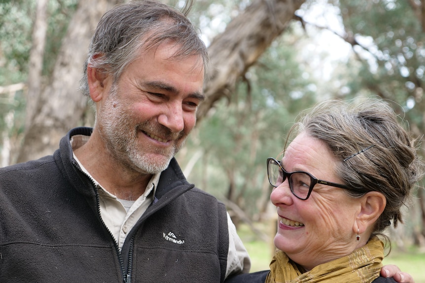 A couple smile while looking into each other's eyes, in front of bushland on their property.