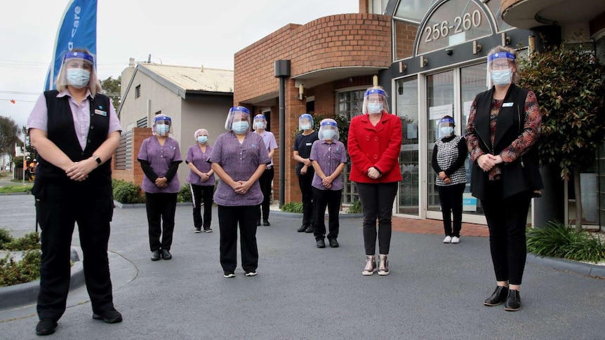 Ten woman stand outside the aged care home in masks and face shields, socially distanced from one another.