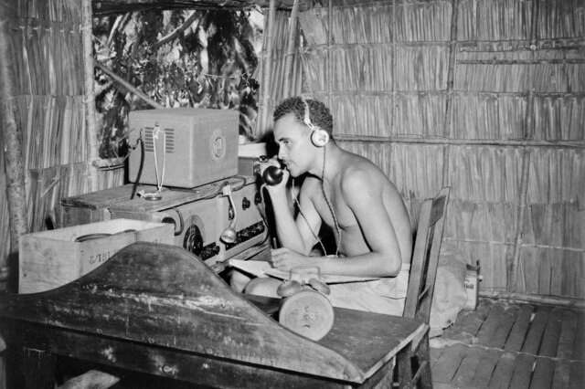 An historical black and white photo shows a man listening into a wireless, in a thatched hut in Solomon Islands.