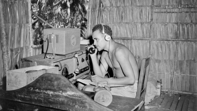 An historical black and white photo shows a man listening into a wireless, in a thatched hut in Solomon Islands.