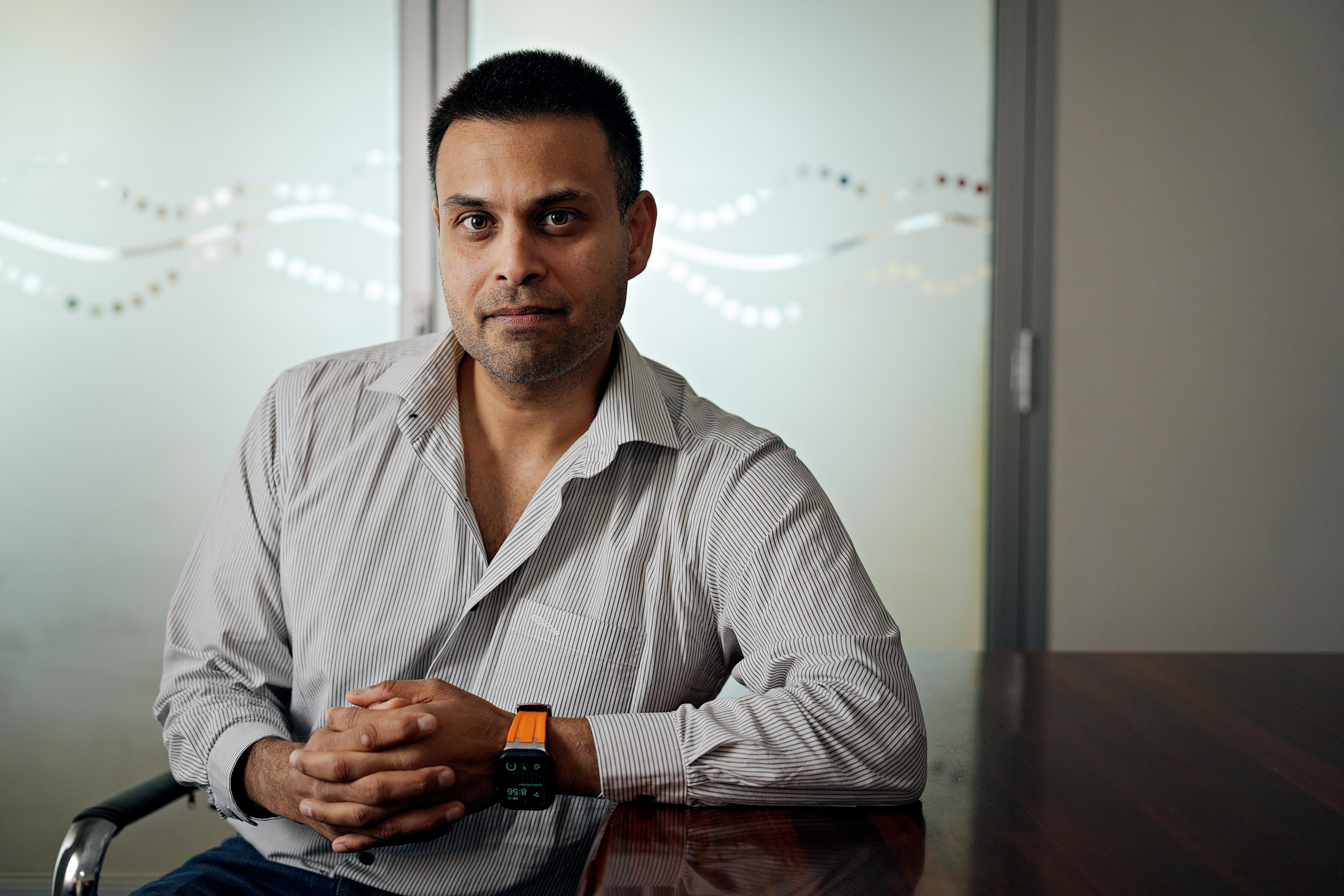 Un hombre con cabello castaño corto y traje de negocios se sienta en una mesa y mira a la cámara. 