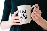 A woman wearing a black jumper holds a mug that reads "Like a boss".