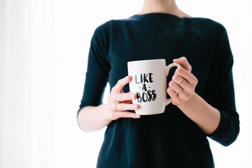 A woman wearing a black jumper holds a mug that reads "Like a boss".