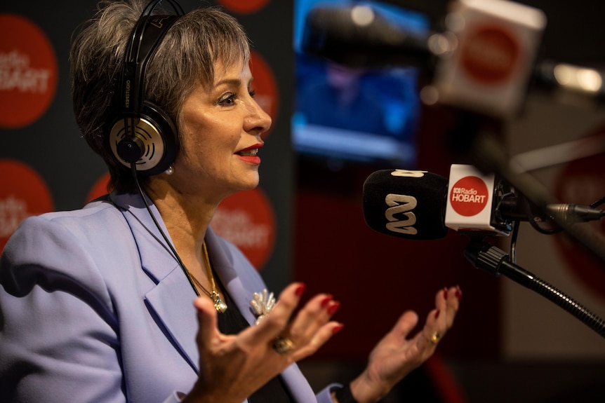 A woman in a purple blazer with headphones on speaking into a micrphone. 