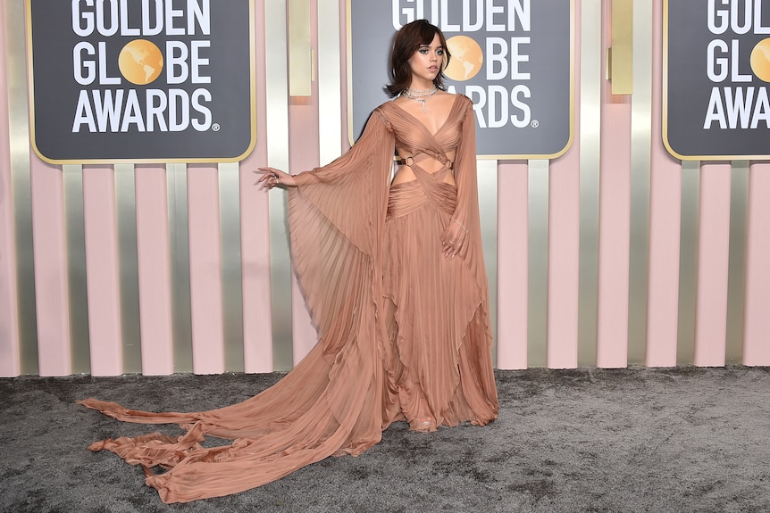 Jenna Ortega wearing alight bronze gown with pleated sleeves, a long train and criss-ross detailing on the bodice. 