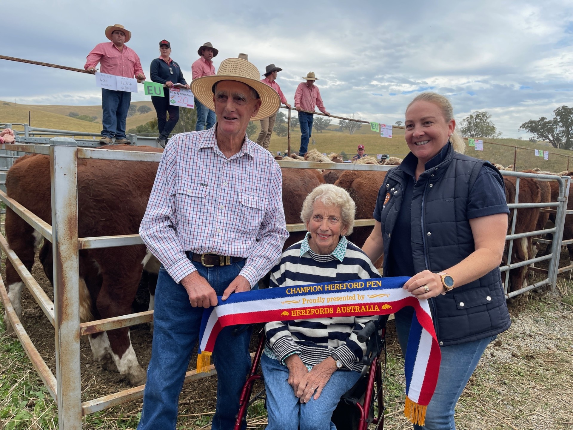 Un hombre y dos mujeres sonríen frente a un establo de ganado en un patio de ventas.