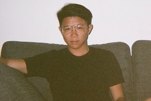 A young Asian man with a neat, trendy hair cut and clear glasses lounges on a grey couch.