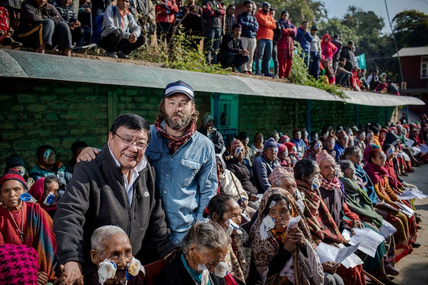 Two men stand in front of a large group of seated Nepalese locals