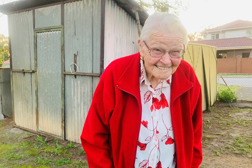 An old woman dressed in red stands in front of a corrugated iron shed