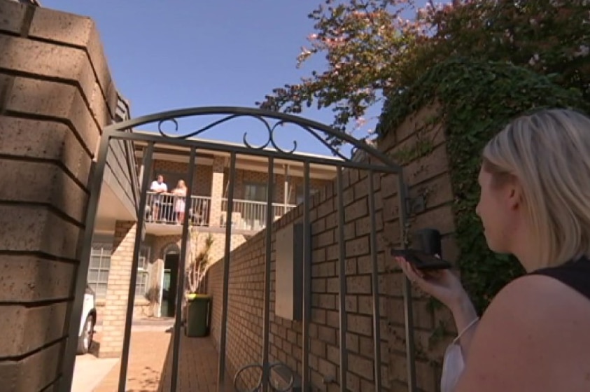 Campbell holding mobile standing behind gate interviewing couple in distance on balcony.