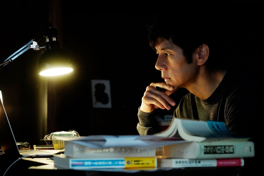 Japanese man wearing olive green shirt looks perplexed while seated at desk illuminated by lamplight.