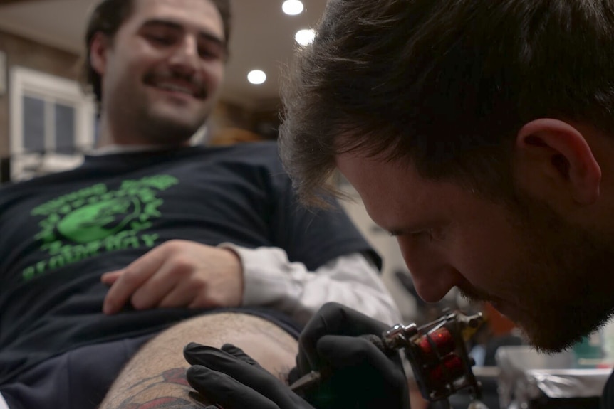 A man focuses on tattooing a leg, the customer smiles in the background. 