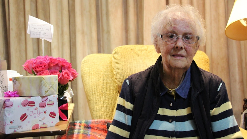 A woman with white hair and glasses looks at the camera from sitting in an armchair, presents at her side