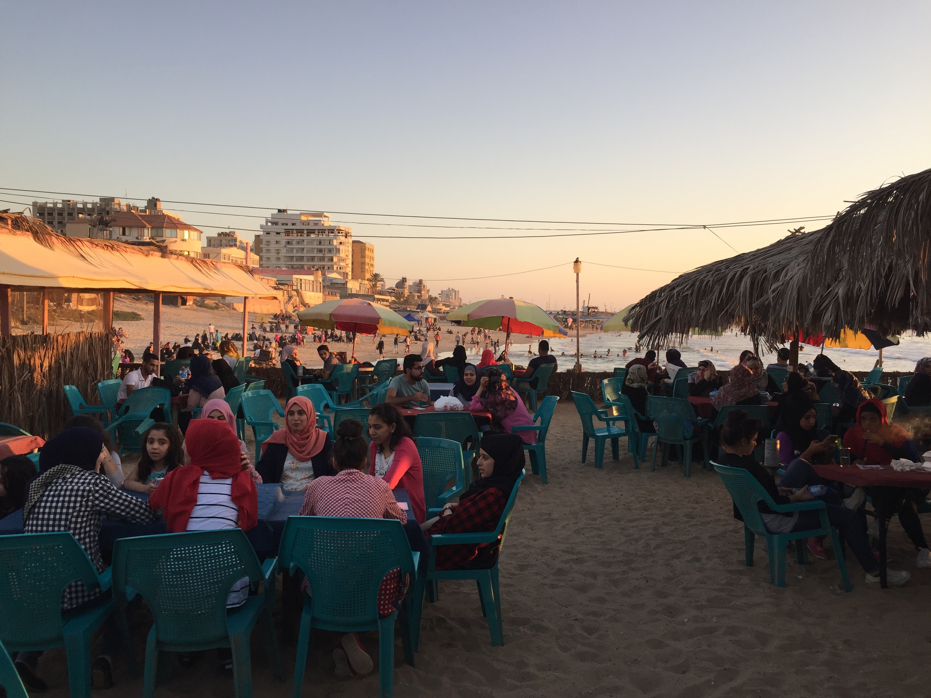 People sit on the beach in Gaza before the war, the tables are on the sand and they are sitting in green plastic chair