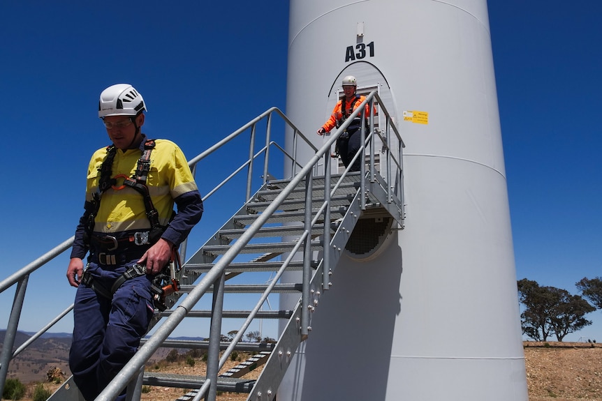 A man and a woman in high vis and helmets walking down metal steps from a door in the base of a wind turbine.