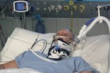 A woman lying in a hospital bed in a coma. She has multiple tubes attached to her and is in a neck brace.
