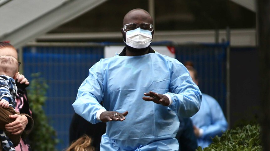 A healthcare worker wearing blue scrubs, safety glasses and a facemask gives directions to people
