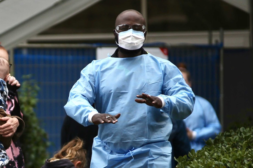 Healthcare worker wearing blue scrubs, safety glasses and a face mask instructs people