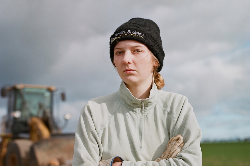 woman stands in grey jumper and black beanie in a field with a tractor in the background