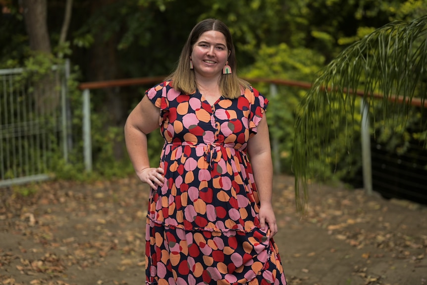 A woman has her hand on her hip, smiling and standing. She wears a coloufrul red and spotty dress. 