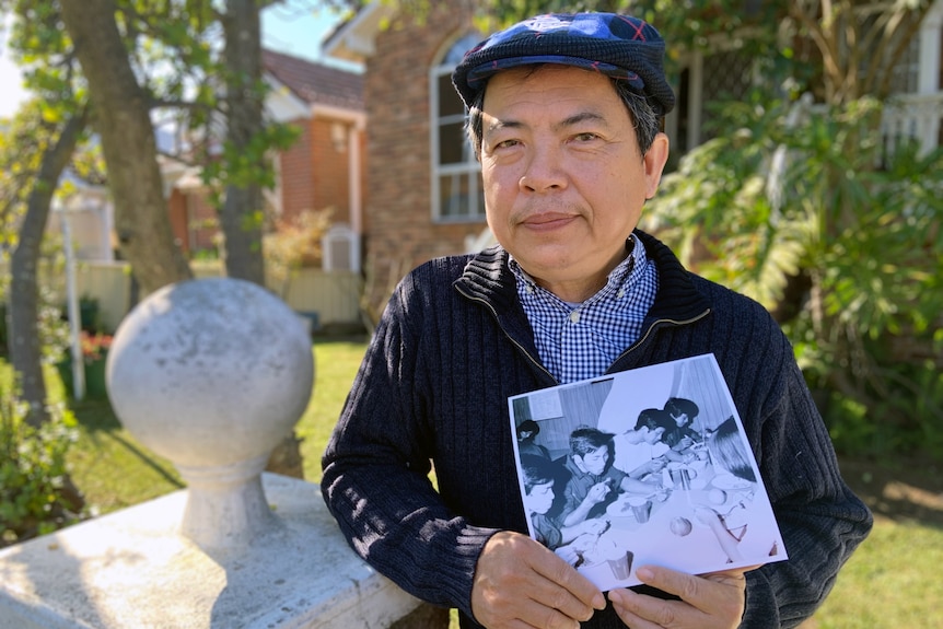 A Vietnamese man in a cap holds a photo of himself about a ship as a refugee
