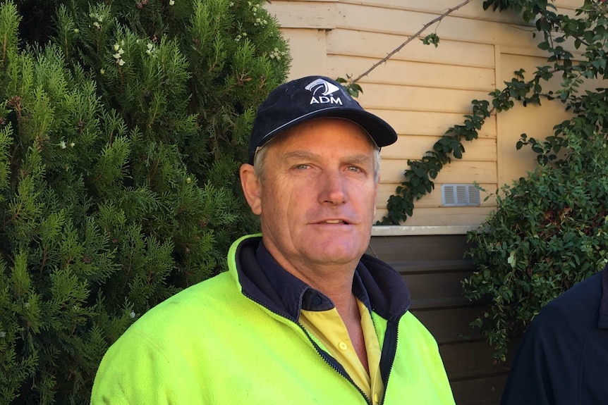 A smiling man wearing a blue cap and a fluro jumper in a garden with trees and a home in the background