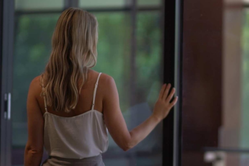 A woman is photographed from behind standing at a window with her hand on the window frame.