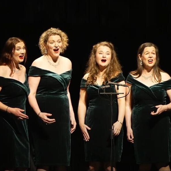 Four women in velvet dresses and glamourous hairdos sing on stage.