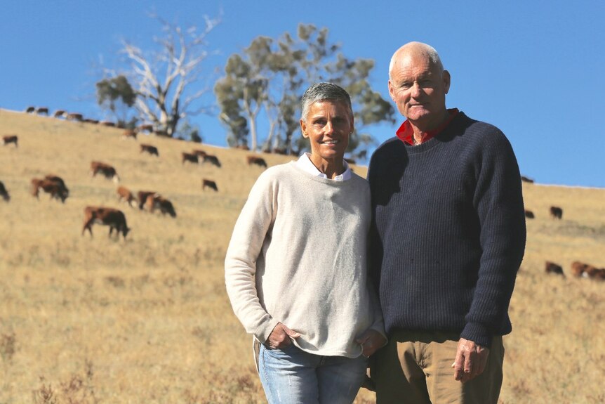 Anna and Michael Coughlan stand in a paddock surrounded by cattle, looking into the camera.
