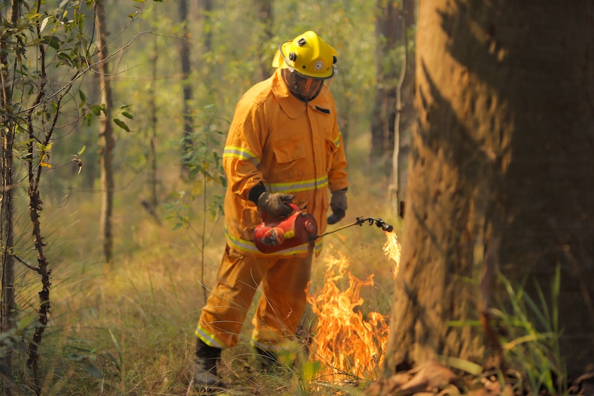 A man in a full body yellow suit and protective gear lights some wild grass on fire. 