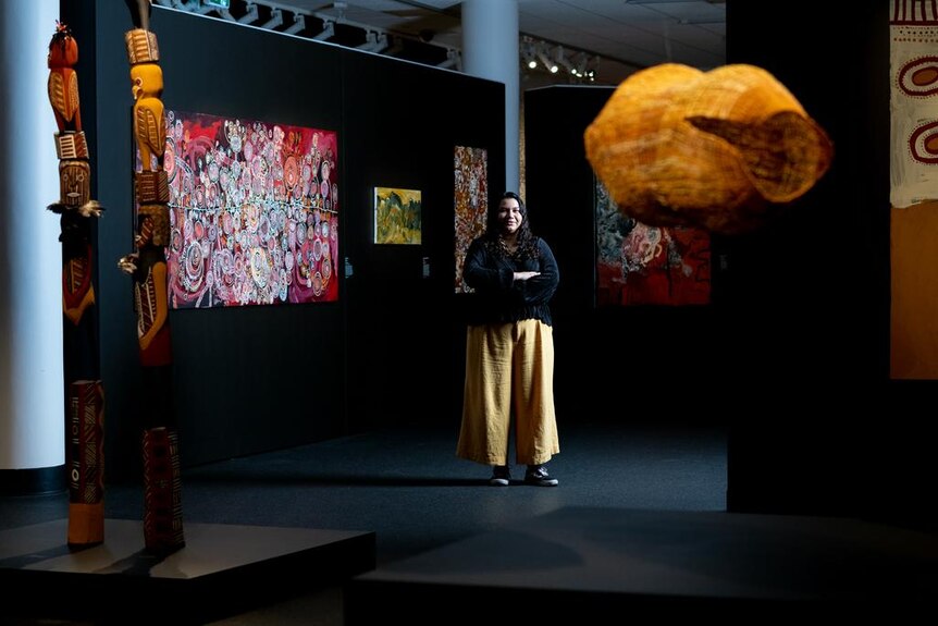 A woman stands in a darkly-lit gallery with out-of-focus, colorful artworks installed around here