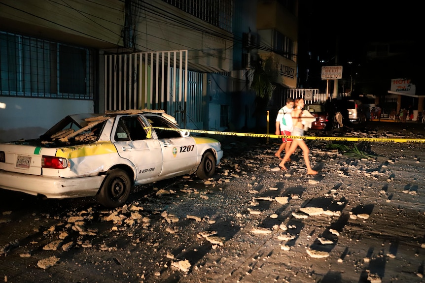 A couple walks past a damaged car and debris.