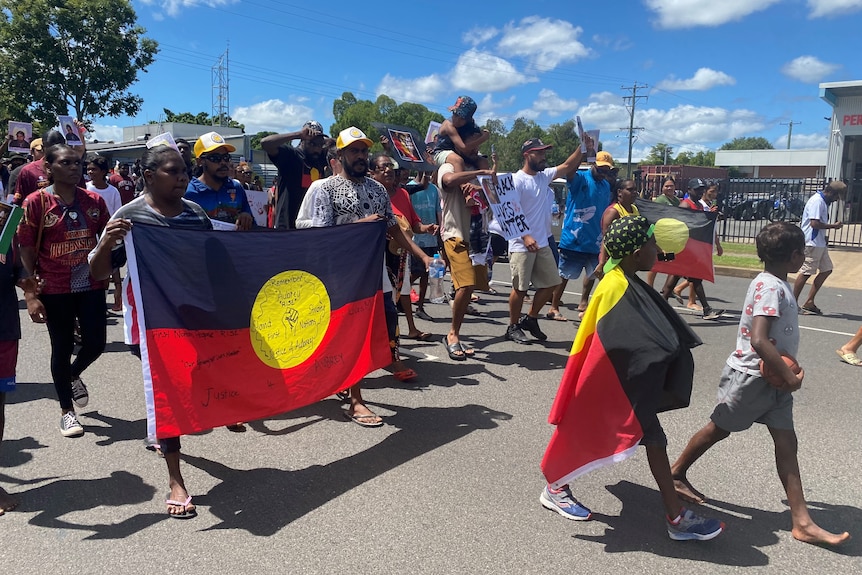 A group of people walk down a road carrying the Aboriginal flag and holding signs reading 'Black Lives Matter'