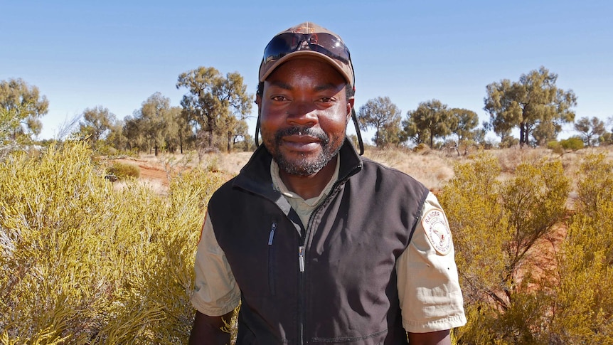 a man in a hat and dark vest next to desert bushes.