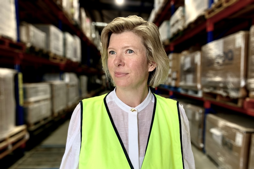Eva Ross looks to her left, wearing a high-vis yellow vest, with rows of pallets behind her.