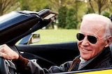 Joe Biden wearing sunglasses and seated behind the wheel of a sportscar