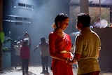 A young woman in a sari talks with head bowed, to a young man as they stand in the spotlight with other players behind.