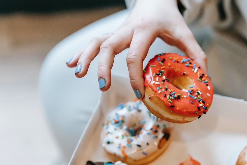 A woman with blue nail polish picks up a pink iced donut from a box of several donuts