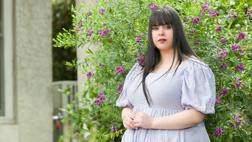 A woman with a neutral expression standing in front of a flowering bush.