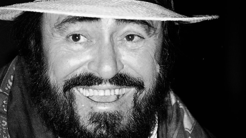 A black and white photograph of Pavarotti looking at the camera wearing a white straw hat and a scarf.
