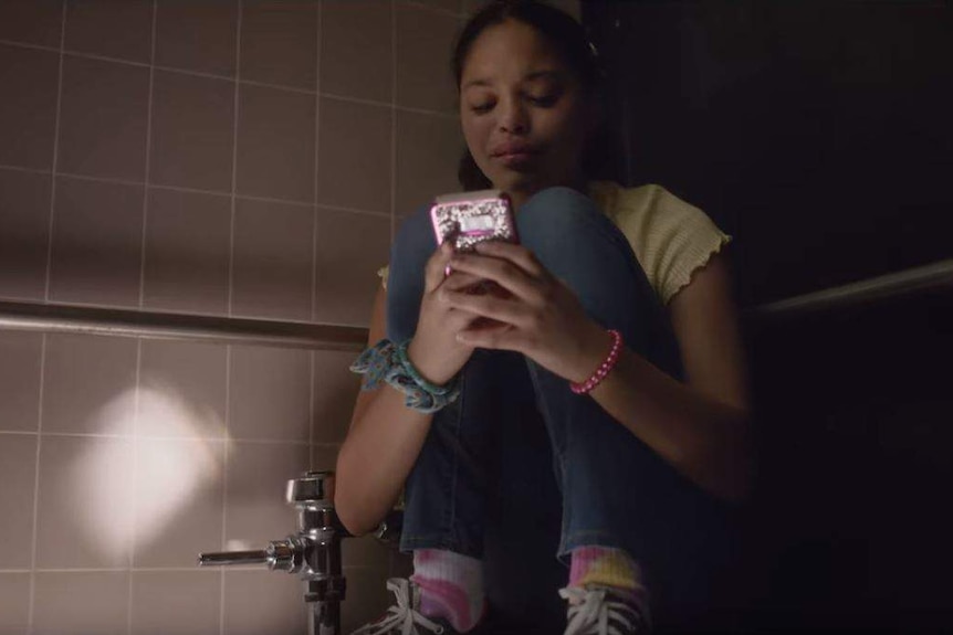 A girl squats on a toilet looking at her phone in a scene from an anti-school-shooting PSA.