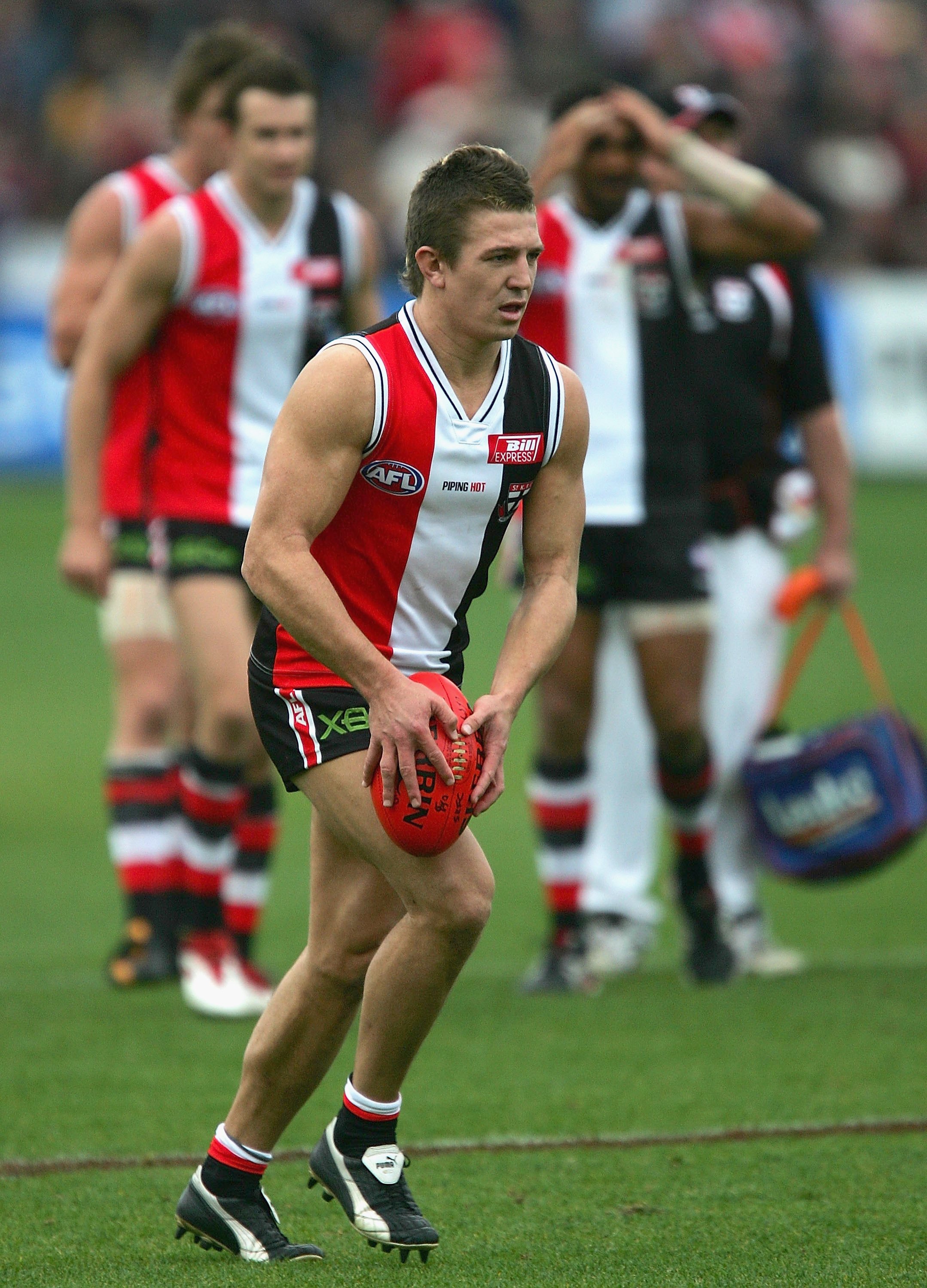 A St Kilda AFL player holds the ball as he prepares to kick for goal at the end of a game, as his teammates watch on.