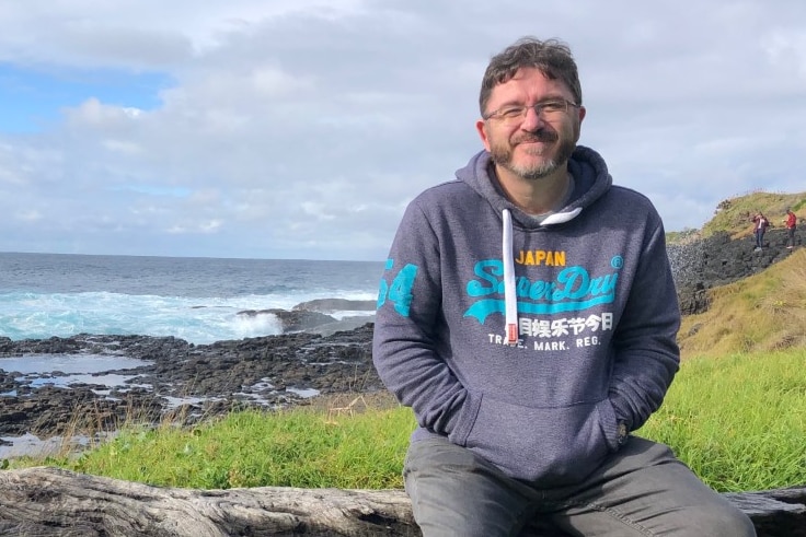 A man wearing a hooded jumper sits on a log in front of a view of grass, rocks and the ocean.