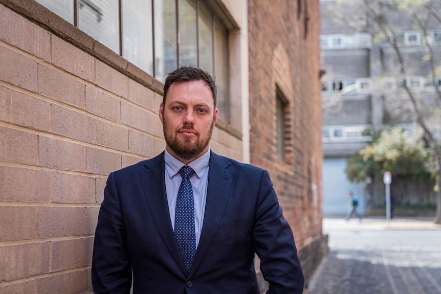 Portrait photo of Brendan Coates. He is wearing a blue suit and tie, has short brown hair, a goatee and brown eyes.